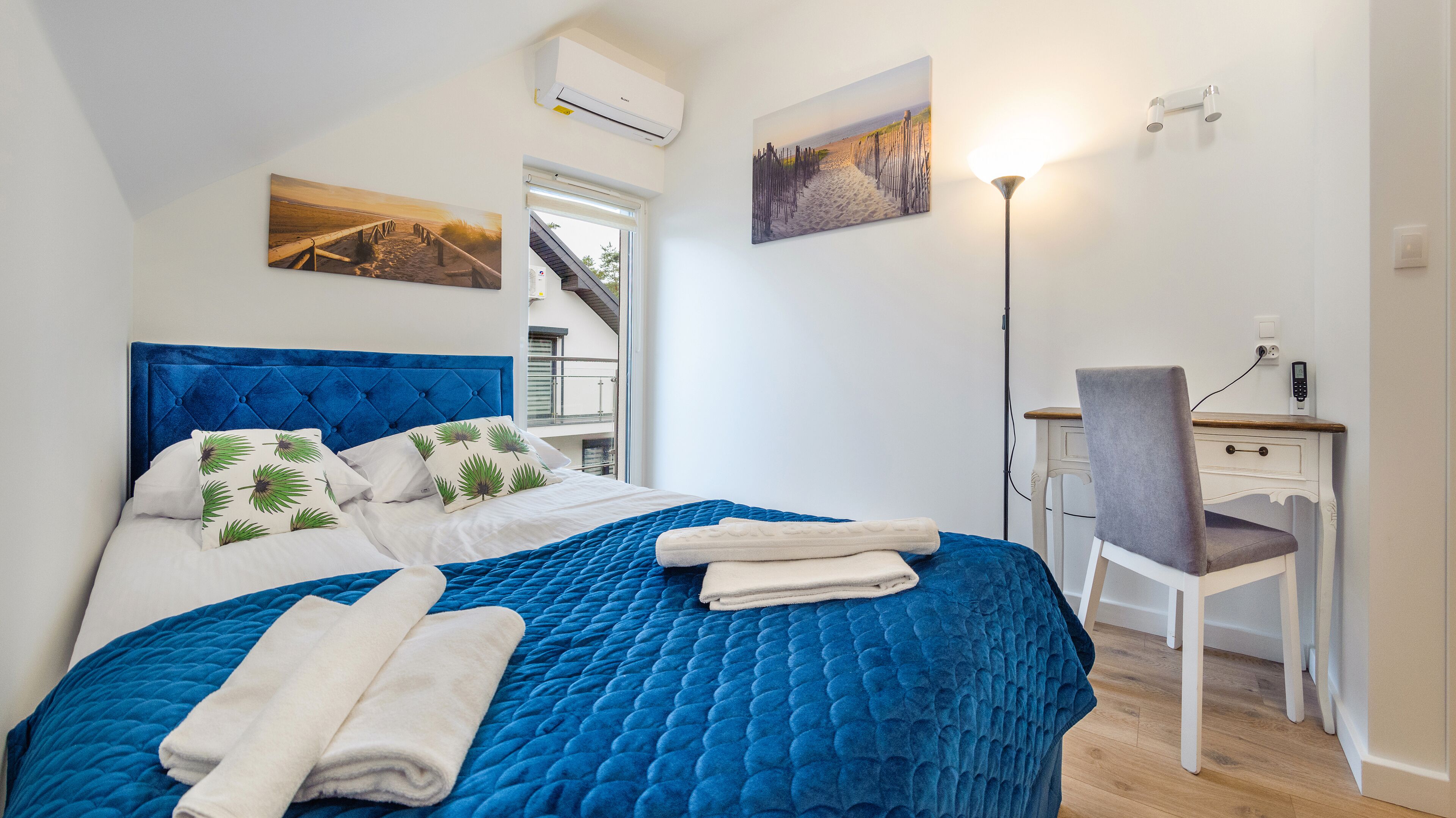 Un dormitorio con una cama grande, un escritorio y una silla. Hay dos cuadros en la pared y un equipo de aire acondicionado sobre la cama.