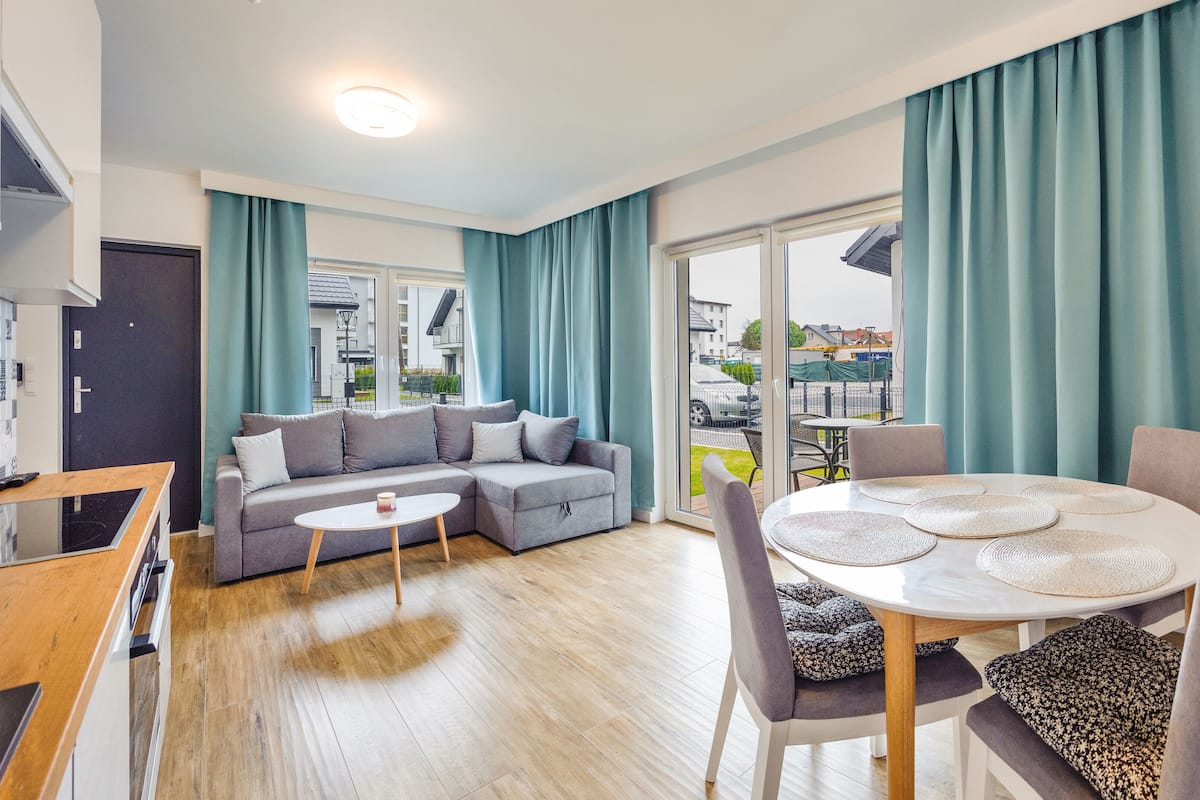 A modern living room with a grey sectional sofa, a round dining table with placemats, and teal curtains.