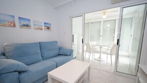 A modern living room with a blue sofa, a white coffee table, and a glass door leading to a dining area with a white table and chairs.