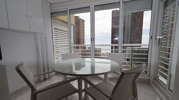 A modern dining area with a glass table and four chairs, large windows with blinds, and a view of tall buildings.