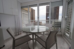 A modern dining area with a glass table and four chairs, large windows with blinds, and a view of tall buildings. - LOS CABALLOS (STUDIO) WITH TERRACE (Benidorm)