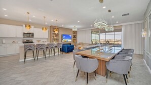 Une cuisine spacieuse avec des meubles blancs, un grand îlot central doté de tabourets, et un coin repas avec une table à plateau en verre et des chaises à revêtement gris.
