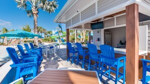 Un espace aménagé au bord de la piscine, avec des chaises et des tables bleues, une terrasse en bois et, en arrière-plan, une vue sur des palmiers et un toboggan aquatique.