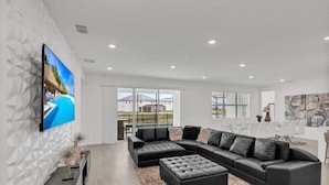 A modern living room with a sectional sofa, a flat-screen TV, and a large window offering a view of the outdoors. - Imagine Your Family Renting This Luxury Contemporary Style Mansion on Veranda Palms Resort Orlando Mansion 5546 (Kissimmee)