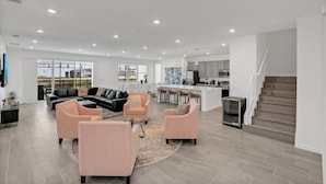 A spacious living room with a large black leather sofa, two pink armchairs, and a coffee table. A kitchen with modern appliances and a bar area is visible in the background. - Imagine Your Family Renting This Luxury Contemporary Style Mansion on Veranda Palms Resort Orlando Mansion 5546 (Kissimmee)