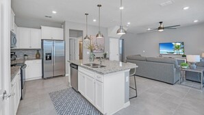 Eine moderne Küche mit Kochinsel, Edelstahlgeräten, Pendelleuchten und einem grautönen Wohnbereich inklusive Sofa und Fernseher.