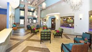 Eine geräumige Lobby mit hoher Decke, einer zentralen Sitzecke mit bunten Stühlen sowie einem Barbereich mit Fernseher und einem großen Fenster, das für natürliches Licht sorgt.