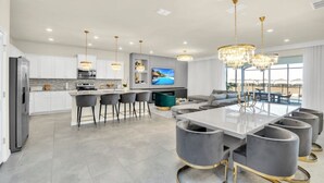 A modern kitchen with white cabinets, a large island with bar stools, a dining area with a glass table and grey chairs, and a living area with a sofa and TV. - Fantastic Mansion on Windsor Island Resort, Orlando Mansion 5660 (Davenport)