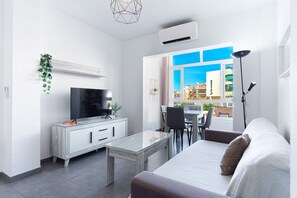 A modern living room with a flat-screen TV, a glass coffee table, a sofa, and a dining area with chairs. - Nevada Centro by Casasol (Nerja)