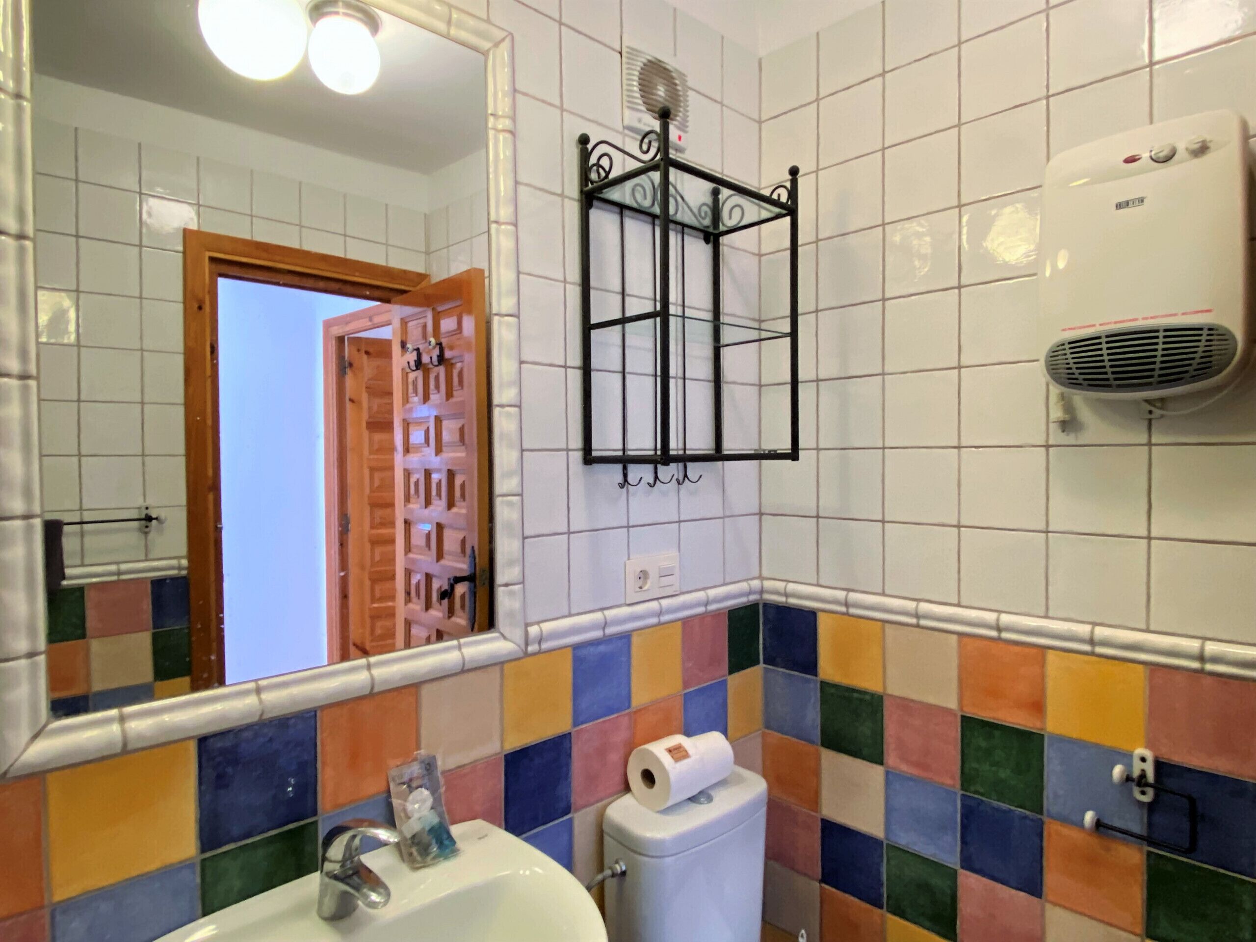 Un baño con pared de azulejos coloridos, un lavabo blanco, un inodoro, un espejo y un secador de pelo en la pared.