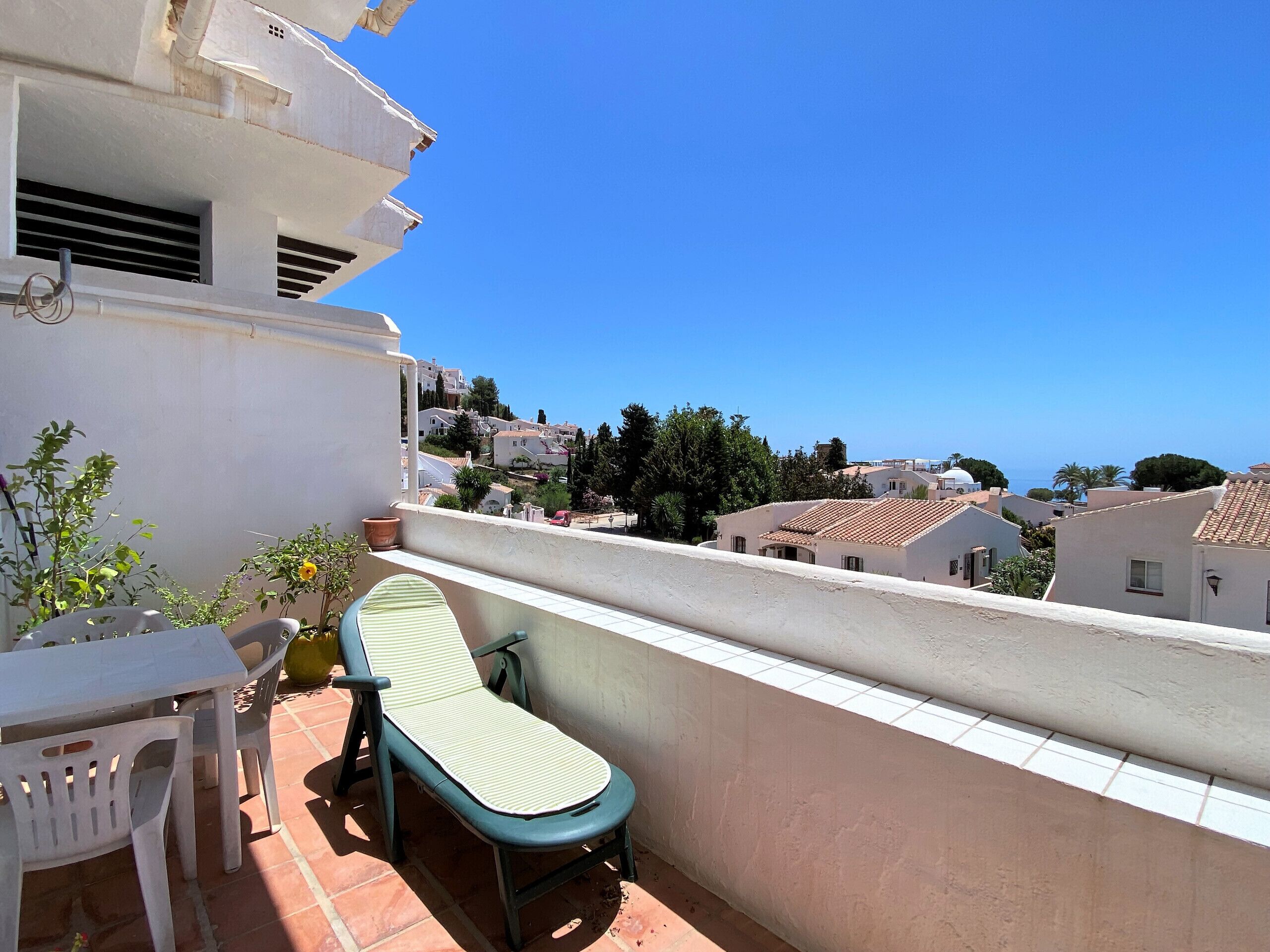 Un balcón con mesa y sillas, una planta en maceta y vistas a una zona residencial con casas y un cielo azul despejado.