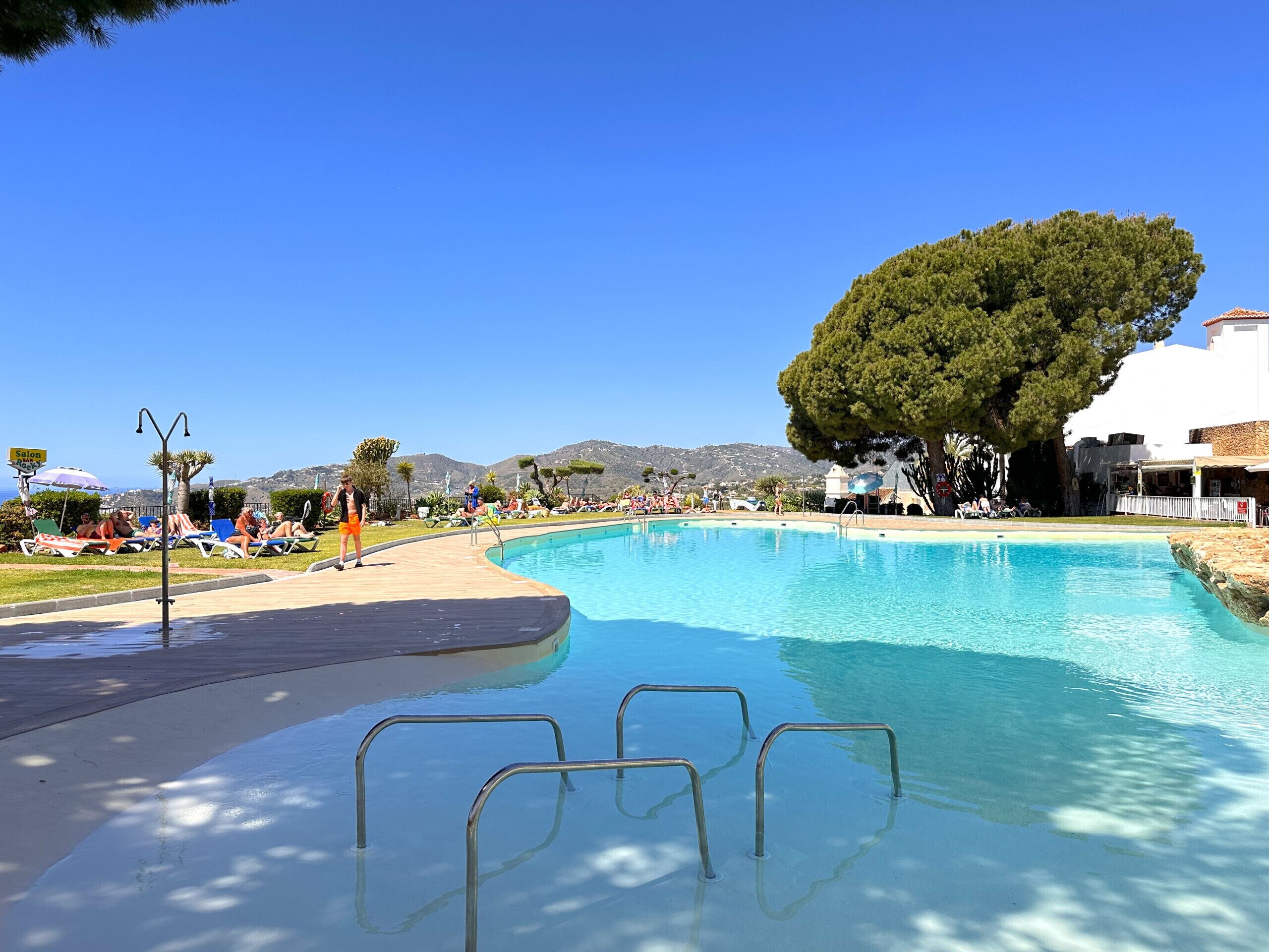 Ein Schwimmbecken mit klarem, blauem Wasser, umgeben von Liegestühlen und Sonnenschirmen, mit einem großen Baum und Bergen im Hintergrund.