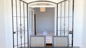 Una habitación con una mesa de vidrio, dos sillas, una cama con sábanas blancas y una puerta con barrotes de metal.