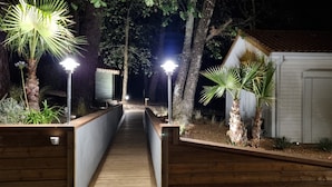 Un sendero de madera que conduce a un edificio con techo de tejas, rodeado de palmeras e iluminado por farolas.