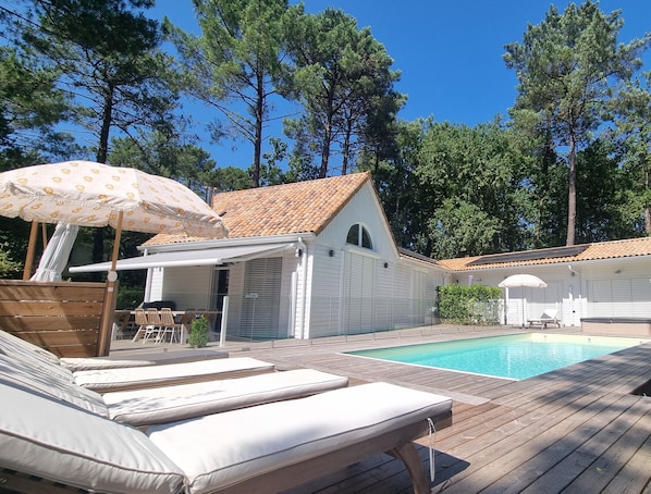 A poolside area with lounge chairs and an umbrella, surrounded by trees and a building with a tiled roof.