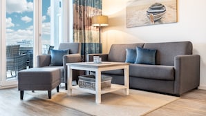 A modern living room with a grey sofa, matching armchair, and a small white table. A large window with a view of buildings and a beach is visible. - Sehr Schönes Ferienapartment mit Meerblick an der Ostsee - nur 25 Meter zum Stra (Großenbrode)