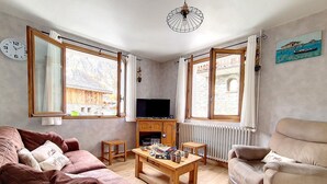 A living room with a sofa, armchair, TV, wooden coffee table, and large windows with curtains. - Family chalet in the center of St Marcel with terrace for 6. (Saint-Martin-de-Belleville)