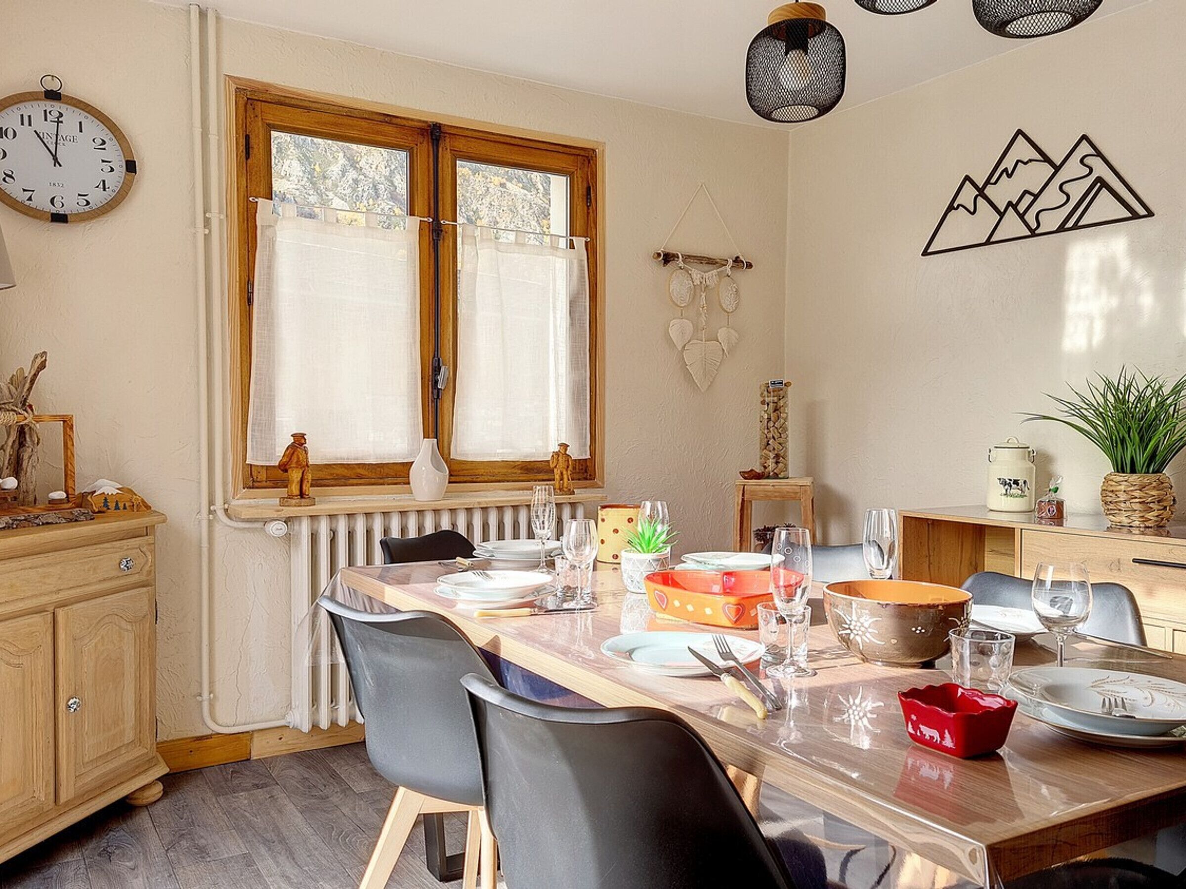 Un coin repas avec une table en bois dressée pour quatre personnes, une fenêtre avec des rideaux, une horloge murale et un élément décoratif fixé au mur.