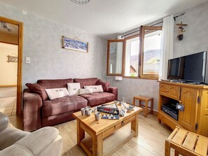 Uma sala de estar com um sofá de couro marrom, uma mesa de centro de madeira, uma TV sobre um armário de madeira e uma janela com vista para o exterior.