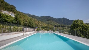 Piscina rectangular con superficie de agua azul cristalina, rodeada por una cerca metálica, con colinas verdes exuberantes y un cielo despejado de fondo.