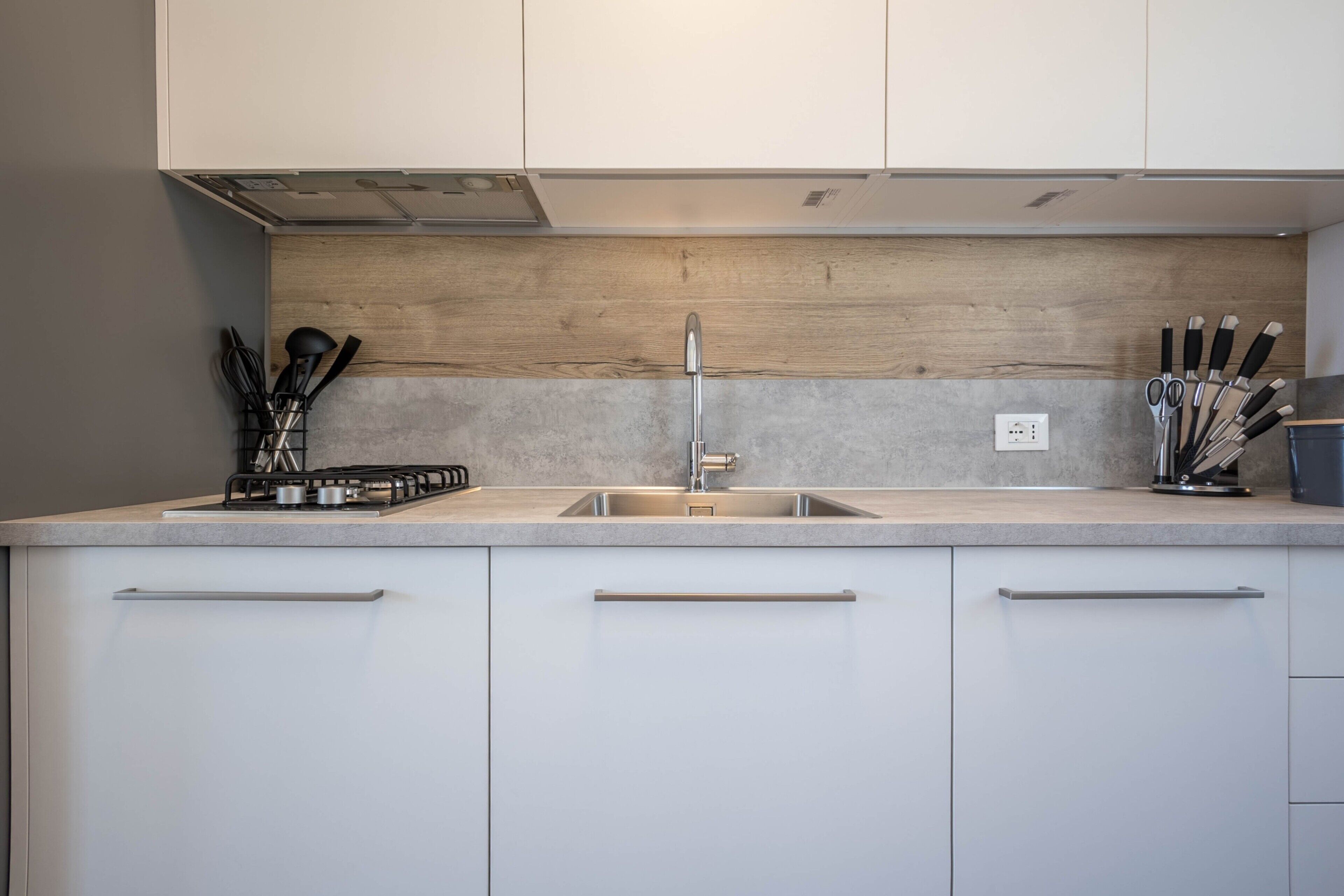 A modern kitchen with a stainless steel sink, wooden backsplash, and white cabinetry.