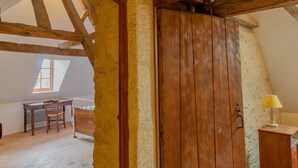 A room with exposed wooden beams, a sloped ceiling, a wooden door, a small table with chairs, and a lamp.