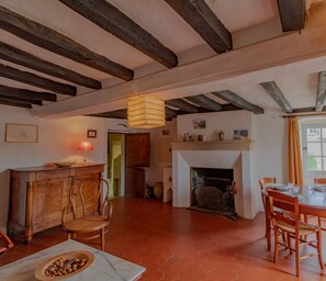 Un interior rústico con vigas de madera a la vista, una chimenea, un comedor con sillas de madera, y una zona de cocina con un armario de madera y un tazón de nueces sobre la mesa.