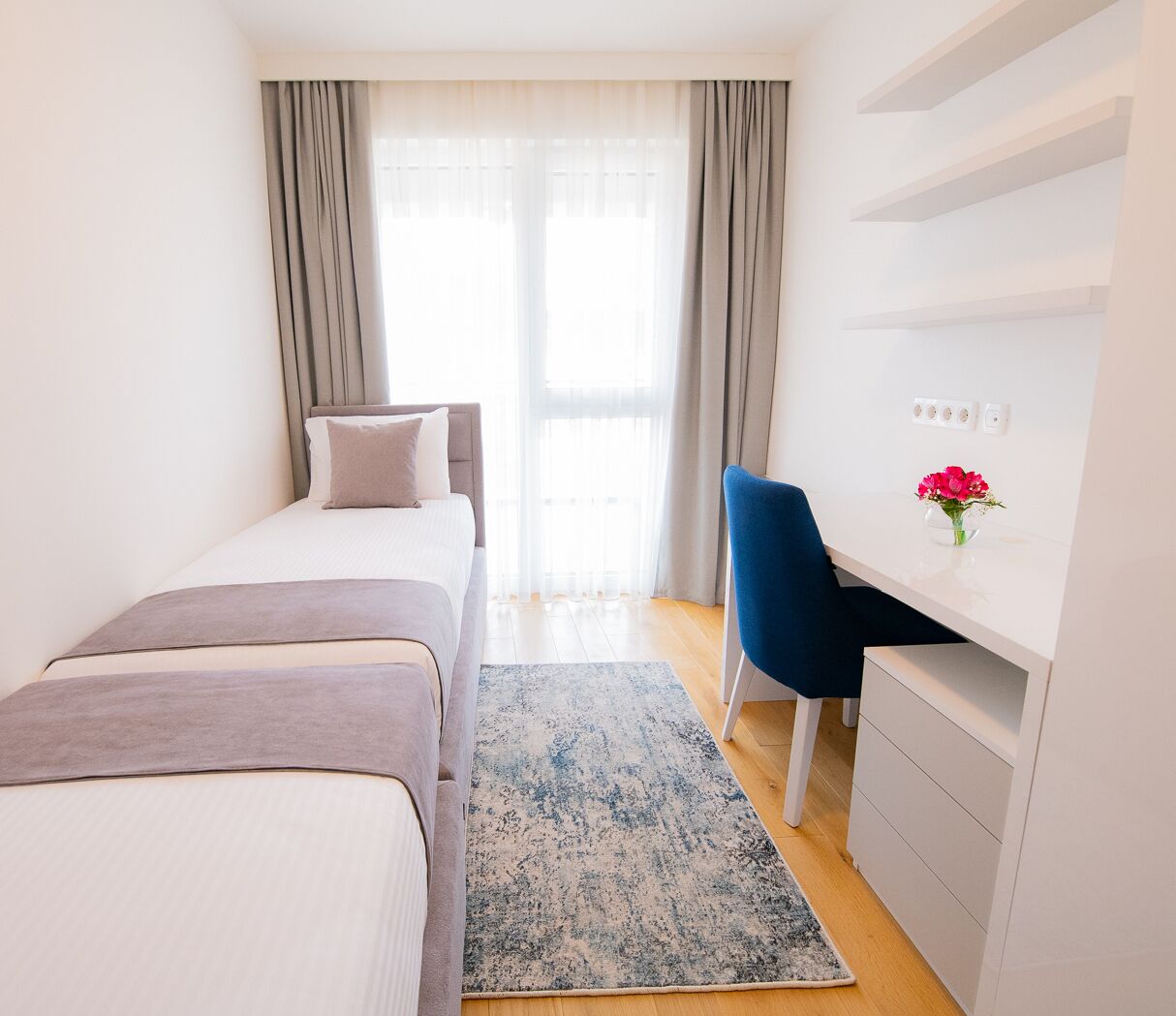 Un dormitorio con cama, escritorio y silla. Hay una ventana con cortinas y un jarrón con flores sobre el escritorio.