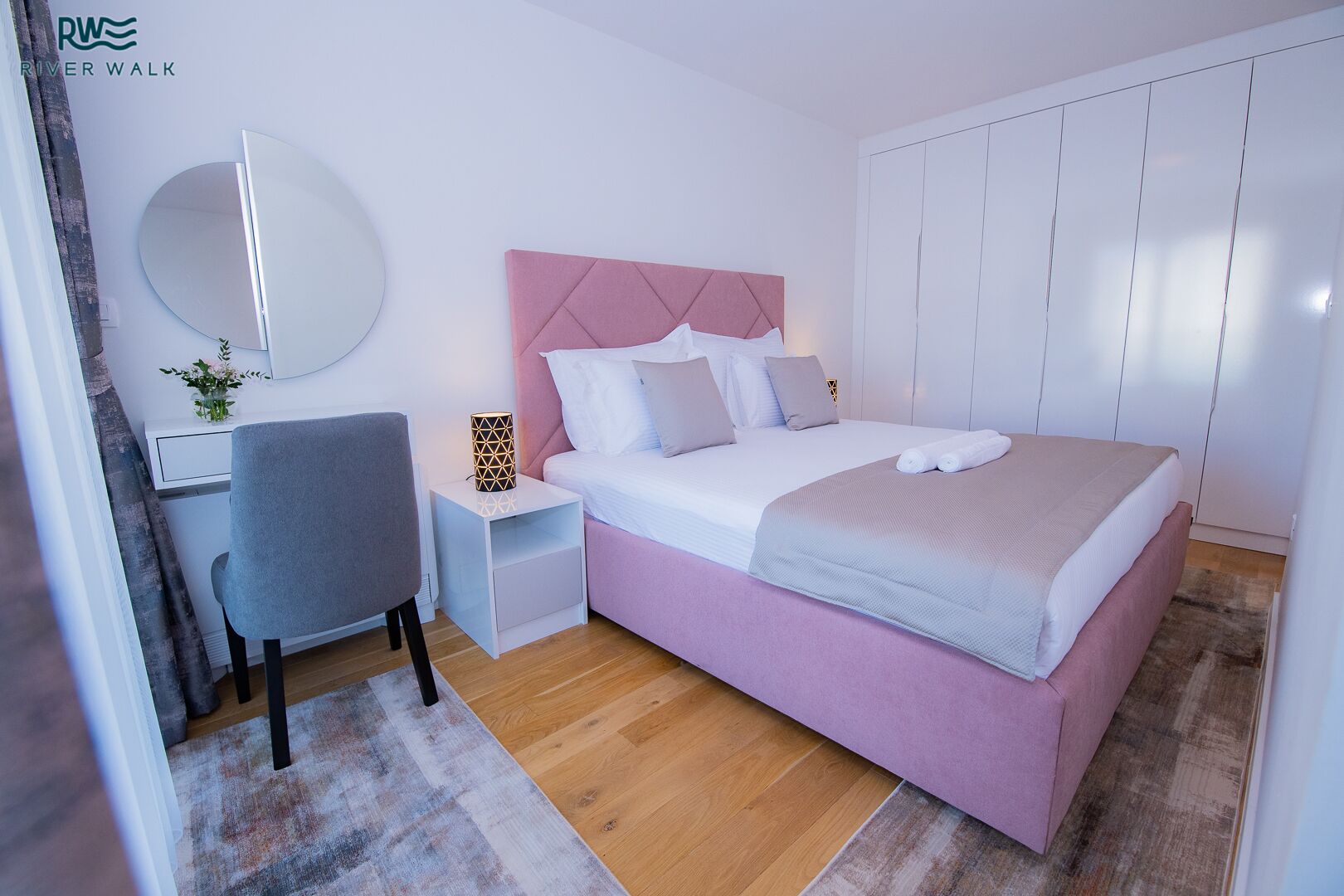 A modern bedroom with a pink bed, white walls, a round mirror, a desk, and a nightstand with a lamp.
