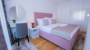 A modern bedroom with a pink bed, white walls, a round mirror, a desk, and a nightstand with a lamp.