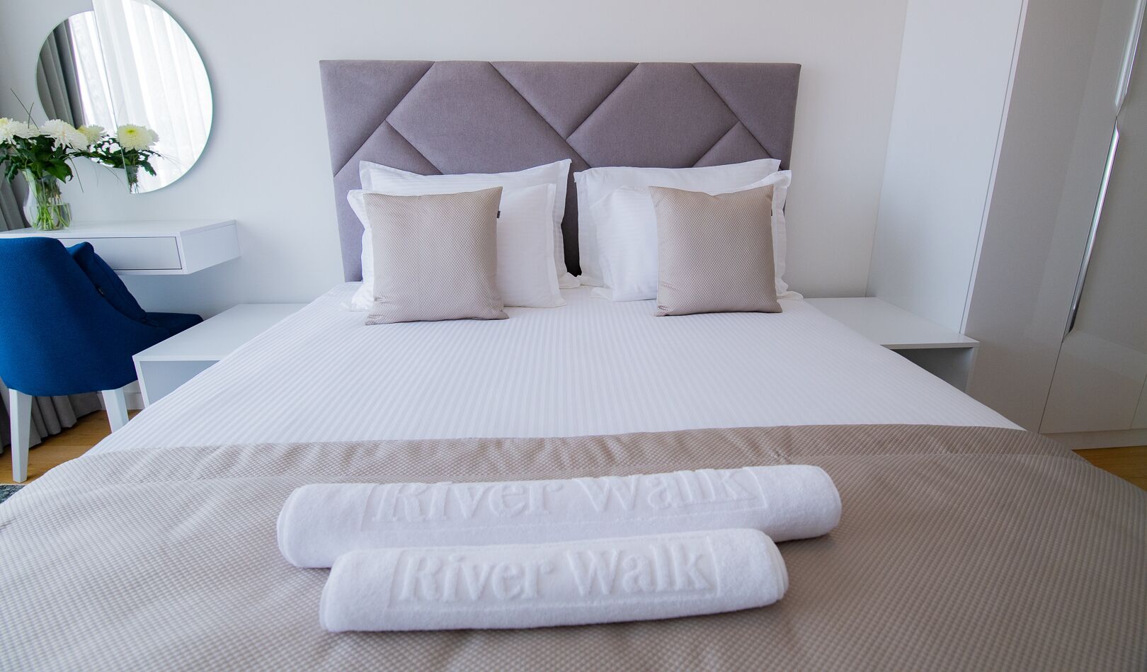 A neatly made bed with a grey upholstered headboard, white bedding, and two rolled towels with "River Walk" branding.