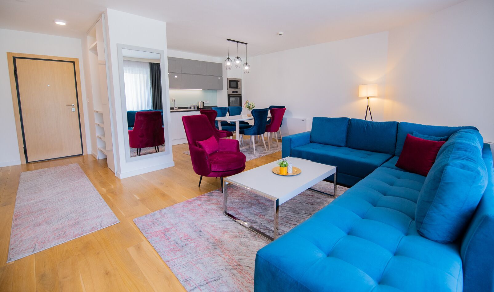 A modern living room with a blue sofa, red and blue chairs, a white coffee table, and a kitchen in the background.