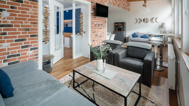 A modern living room with a brick wall, a grey sofa, a coffee table, and a dining area with a white table and grey chairs. - The sea always works - Winterstrasse 23 (Norderney)