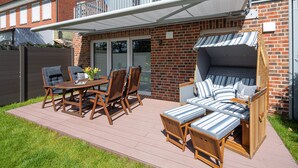 A patio with a wooden table and chairs, a wicker sofa, and a striped cushion set against a brick wall. - // VACATION APARTMENT CLOUDLESS // (Norderney)