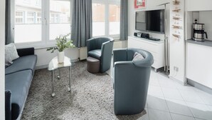 A modern living room with a grey sofa, two armchairs, a glass coffee table, and a TV. - // REGINA MARIS - APARTMENT 1 // (Norderney)