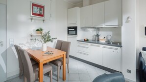 A modern kitchen with white cabinets, a built-in oven, and a dishwasher. There is a dining table with chairs and a vase with flowers. - // REGINA MARIS - APARTMENT 1 // (Norderney)