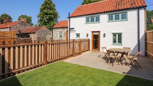 Une maison blanche à deux étages, avec un toit recouvert de tuiles rouges, une clôture en bois et une terrasse équipée d’une table et de chaises.