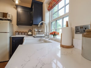 A kitchen with dark cabinets, a white marble countertop, a sink, a kettle, and a window with a view of greenery. - Postbox Cottage (Thetford)