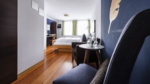 A modern hotel room with a wooden floor, a bed with a white headboard, a small round table with a lamp, and a blue wall with a partial face mural. - Hotel Bären (Marbach am Neckar)