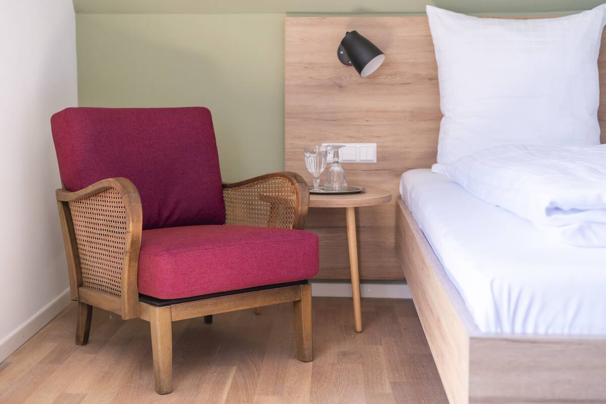 A room with a wooden bed, a red upholstered chair, a small wooden table with a lamp, and a wall-mounted switch.