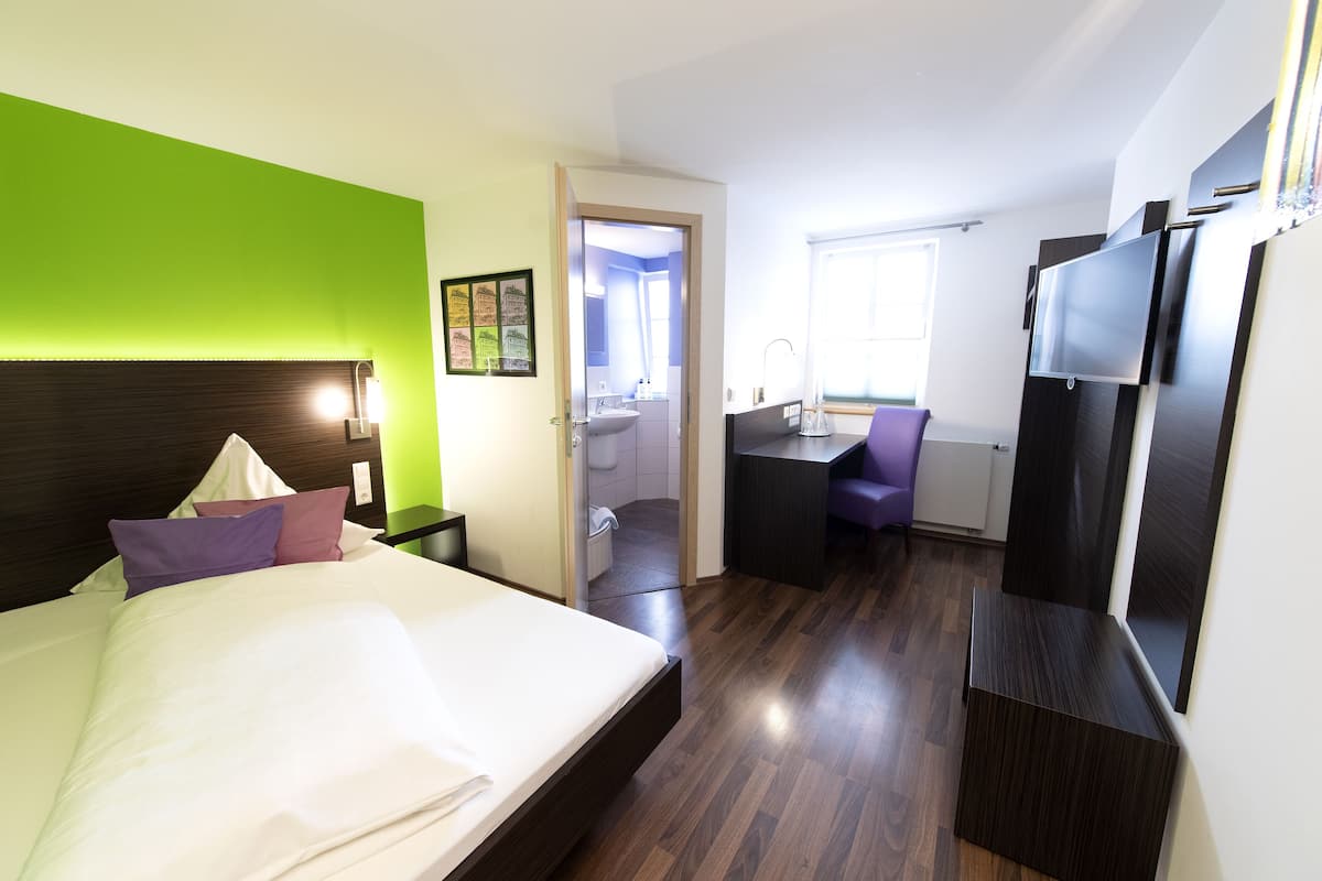 A hotel room with a bed, a desk, a TV, and a bathroom visible through an open door.