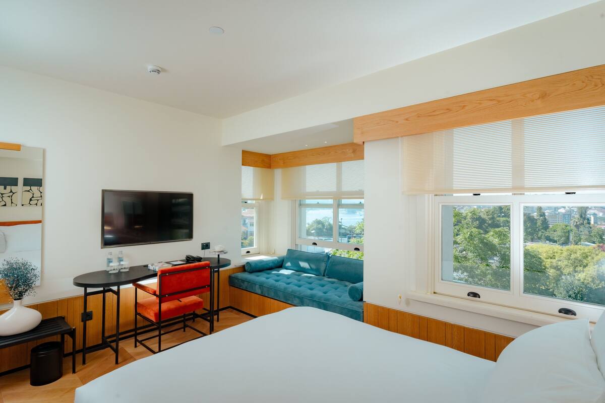 A modern hotel room with a bed, a seating area with a blue sofa, a small table with a chair, a TV, and a window with a city view.