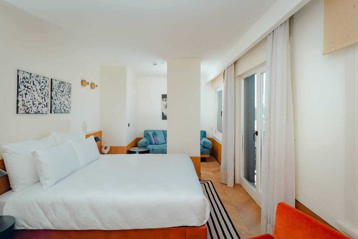 A hotel room with a large bed, a small seating area with a blue chair, and a view of the city through the window.
