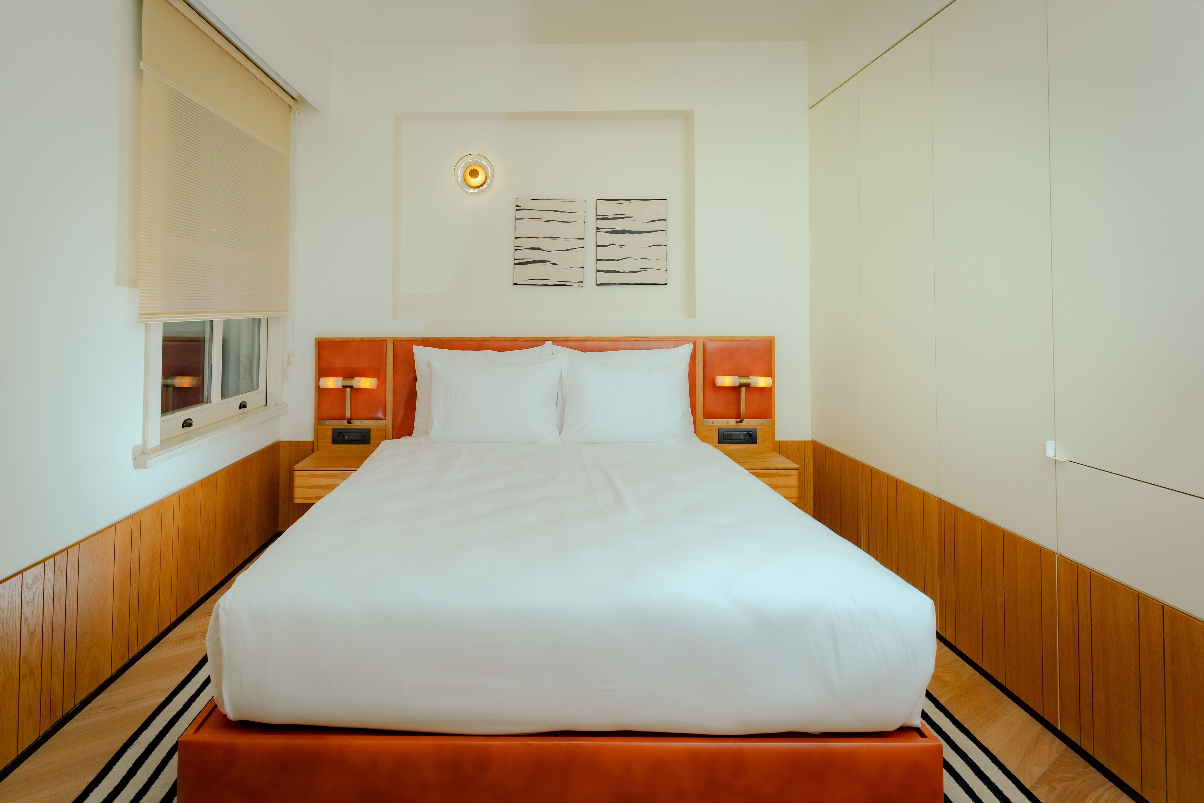 Een net opgemaakt bed met witte lakens, twee nachtkastjes met lampen, een raam met zonwering en twee abstracte kunstwerken aan de muur.
