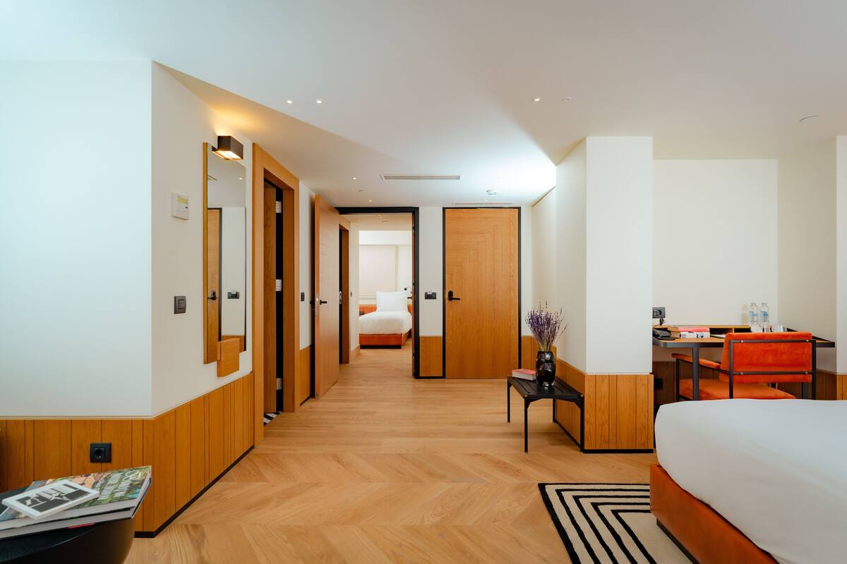 A hotel room with a bed, desk, chair, and a small table with a vase. There is a mirror on the wall and a door leading to another room.