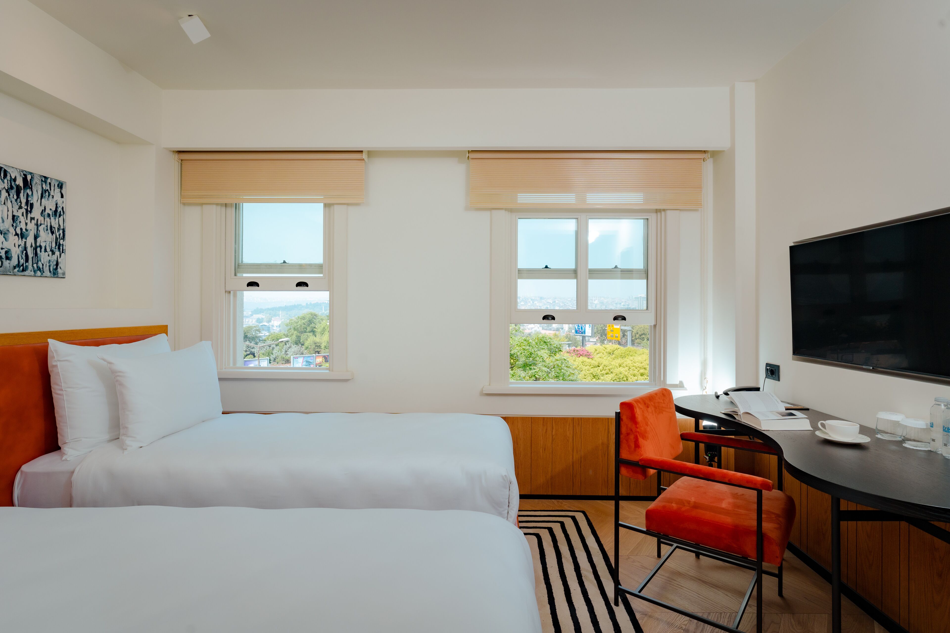 A hotel room with two beds, a desk with a chair, a TV, and large windows with blinds.