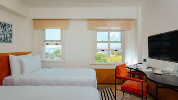 A hotel room with two beds, a desk with a chair, a TV, and large windows with blinds.