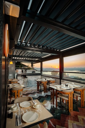 A restaurant with tables set for dining, a bar area, and a view of the sunset over a body of water.