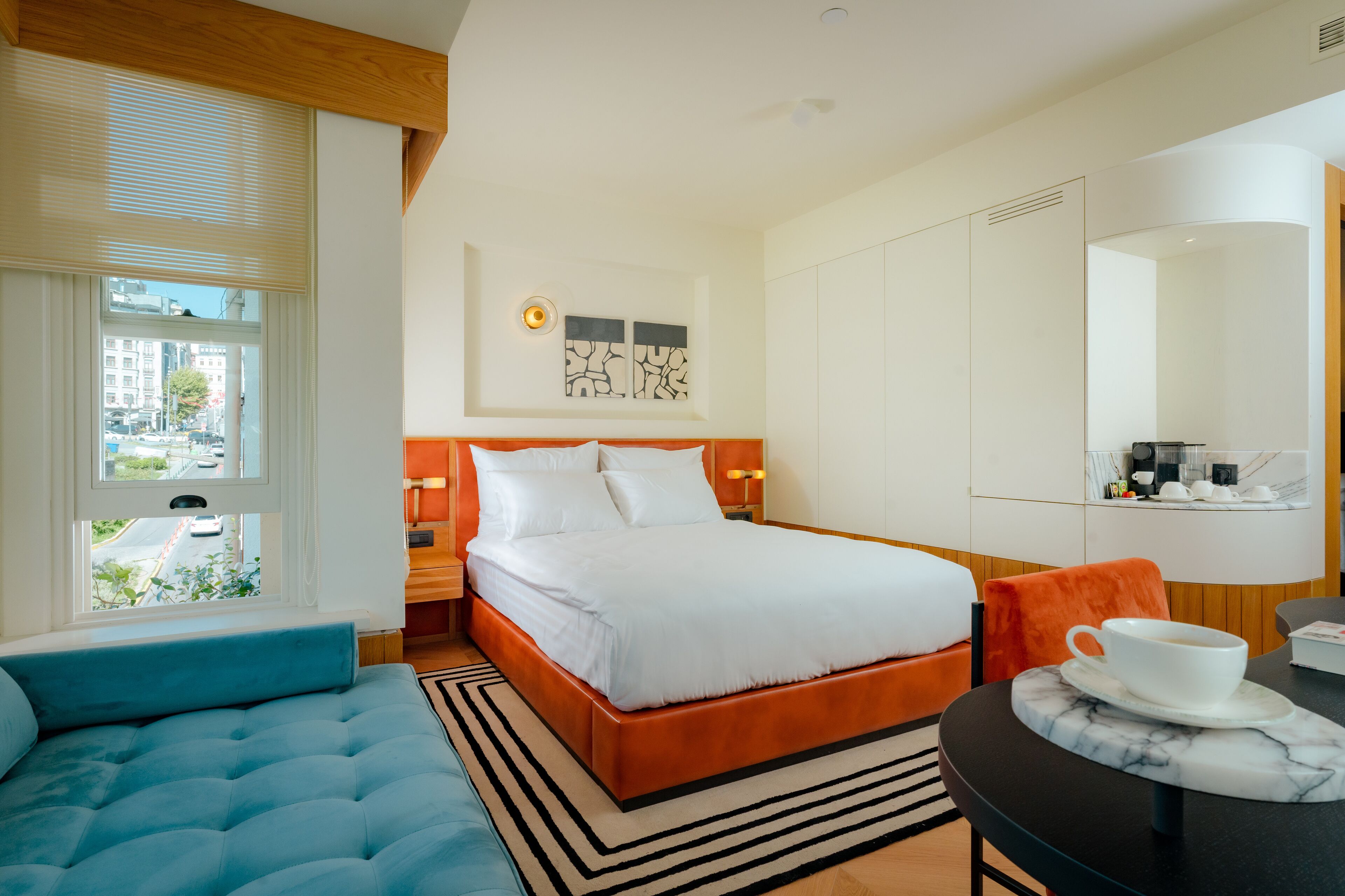 A modern hotel room with a large bed, a seating area, a dining table, and a view of the city through the window.