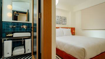 A hotel room with a bed, a bathroom with a mirror, and a shelf with towels.